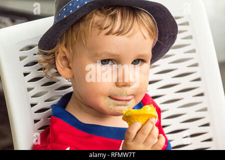 Un portrait d'un charmant garçon avec un chapeau bleu profitant d'une délicieuse crème glacée. Il est titulaire d'un cornet de crème glacée jaune dans sa main. Banque D'Images