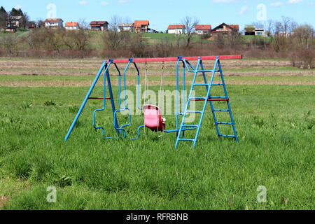 L'équipement de jeu public extérieur avec balançoire métal bleu et du châssis du siège en plastique délabrée entourée d'herbe non coupée et sur le terrain avec la famille Banque D'Images