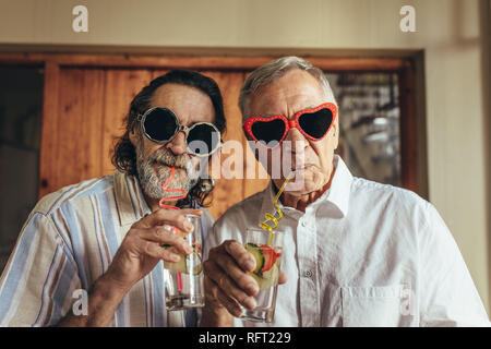 Les hommes portant des lunettes de soleil drôle boire du jus avec de la paille. Personnes âgées amis avec lunettes folles buvez du jus à l'intérieur. Banque D'Images