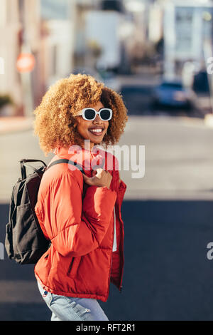 Femme afro-américain en vacances, marcher autour de la ville. Smiling tourist femme portant des lunettes de soleil et un sac à dos marche sur rue. Banque D'Images