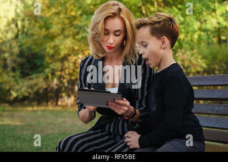 Young attractive la mère et le fils sont assis sur un banc dans le parc de la ville. Femme est montrant quelque chose sur l'écran du PC tablette. Boy est très surpris. Temps de la famille Banque D'Images