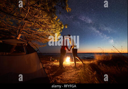 Nuit de camping bleu en mer sous ciel étoilé. Jeune couple, homme et femme s'embrasser au camp. L'eau claire et les lumières de la ville sur la rive lointaine sur backgroun Banque D'Images