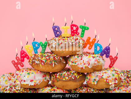 Le chocolat et les beignets blanc gelé recouvert de candy sprinkles empilées en tas avec gâteau Joyeux Anniversaire bougies vivement. Fond rose. Banque D'Images