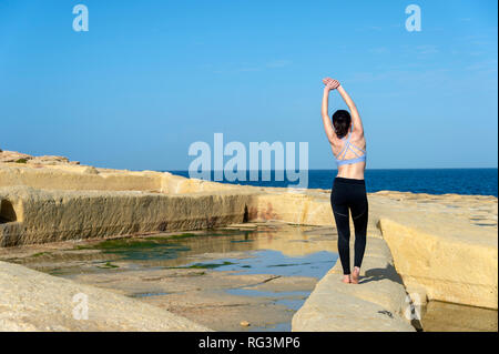 Vue arrière d'une femme faisant un côté un étirements à l'extérieur par la mer le port d'un soutien-gorge de sport et leggings Banque D'Images