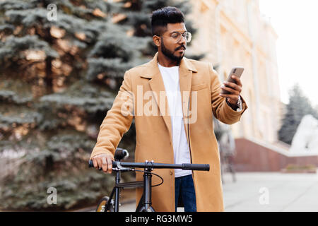 Bel homme barbu en costume classique est à l'aide d'un téléphone intelligent et souriant tout en vélo en ville