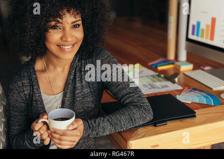 Portrait of happy young woman having coffee at home office 24. Femme africaine assise à son bureau avec une tasse de café à la caméra et au sourire. Banque D'Images