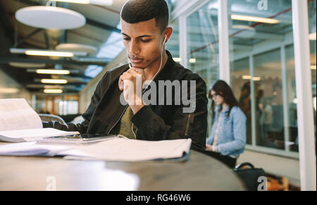 Man wearing earphones looking at digital tablet while sitting at university library. Male student studying at college campus using digital tablet. Banque D'Images