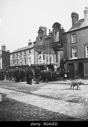 Une petite foule rassemblée sur une rue pavée à l'extérieur d'un bâtiment endommagé d'incendie, peut-être en Ecosse. Vers 1915 Banque D'Images