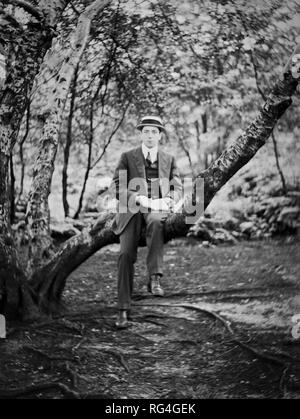 Un jeune homme portant un chapeau en paille assis sur une branche d'arbre avec un livre ouvert sur son genou, vers 1920 Banque D'Images