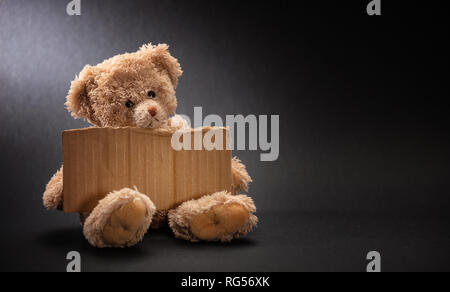 Les pauvres sans-abri la mendicité des enfants. Ours triste, holding a blank sign en carton, assis sur fond noir, copy space Banque D'Images