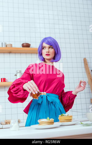 Portrait de femme au foyer avec des cheveux violets et sirop de vêtements colorés sur les crêpes dans la cuisine Banque D'Images