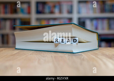 Dés dans un livre forment le mot 'read'. Livre est couchée sur un bureau en bois en face d'une étagère. Banque D'Images