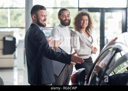 Les clients heureux, debout près de marchand de voiture et souriant. Les hommes et belle femme à la recherche de l'automobile, de l'observation. Manager en veste bleu foncé du dossier, montrant des véhicules pour les clients. Banque D'Images