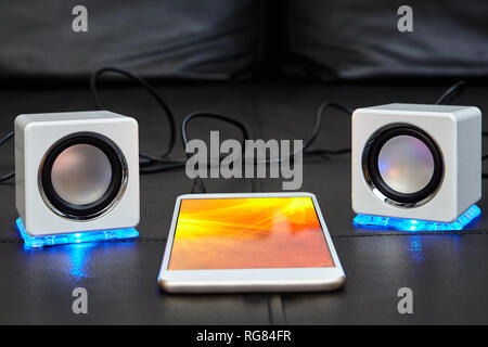 Gadgets modernes se trouvent sur le siège d'un canapé en cuir noir, un smartphone avec un écran d'affichage de couleurs vives et deux mini haut-parleurs avec éclairage LED blu Banque D'Images