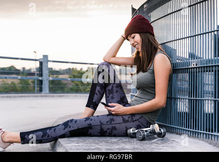 Jeune femme assise sur le sol après l'entraînement des haltères courts, using smartphone Banque D'Images