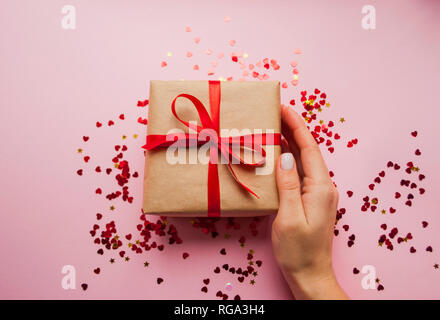 Boîte-cadeau enveloppé dans du papier craft de couleur brune et à égalité avec l'arc rouge sur fond rose avec confetti rouge. Banque D'Images