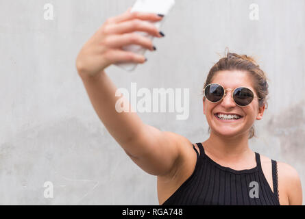 Portrait of laughing young woman wearing sunglasses en tenant avec smartphone selfies Banque D'Images