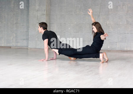 Jeune couple équilibre acrobatique pratiques. Les jeunes danseurs de style de mode portant des costumes noirs sur fond gris. Banque D'Images