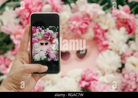 Blogging Instagram concept. Main tenant le téléphone et la prise de photo de girly élégant télévision laïcs de forme de coeur avec des lunettes roses pivoines rose et blanc o Banque D'Images