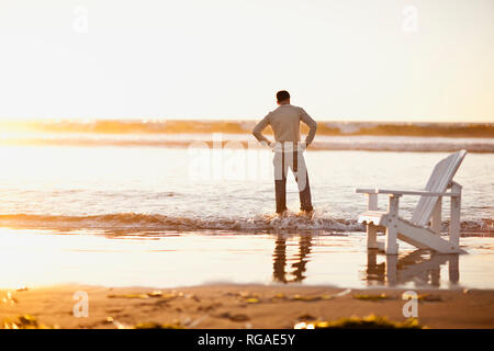 L'homme avec sa main sur ses hanches patauge dans la mer et regarde l'eau éclaboussant son jean au coucher du soleil. Banque D'Images