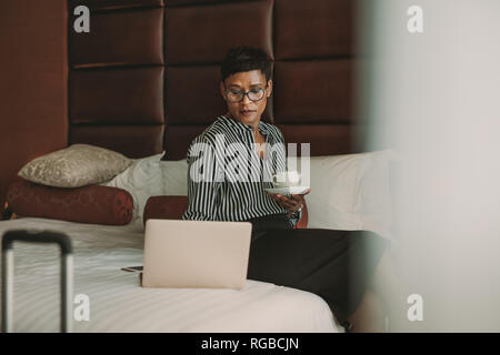 Femme africaine en vêtements officiels assis sur le lit avec du café dans l'hôtel, à l'aide de l'ordinateur portable. Construction à partir de la chambre d'hôtel. Banque D'Images