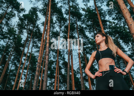 Belle fille femme athlète, se tient en arrière-plan des forêts d'arbres, l'air frais. Mode de vie actif dans le parc l'été. Sportswear décontracté. En mains et téléphone Banque D'Images