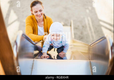 Baby Girl climbing sur shute sur l'aire de jeux étant détenu par sa mère Banque D'Images