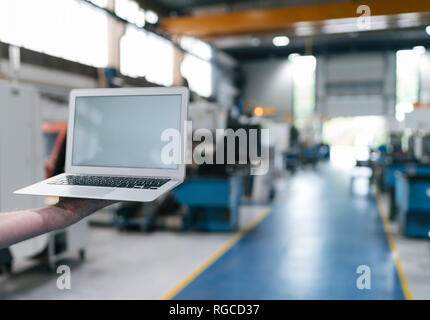 Main tenant un ordinateur portable avec écran vide dans un atelier d'usine Banque D'Images