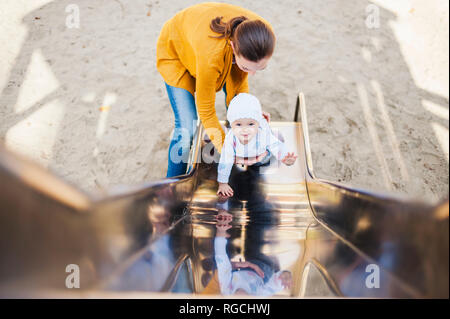 Smiling baby girl sur shute sur l'aire de jeux étant détenu par sa mère Banque D'Images