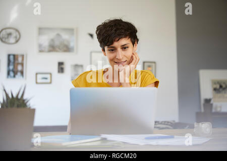 Portrait of smiling woman at home sitting at table using laptop Banque D'Images