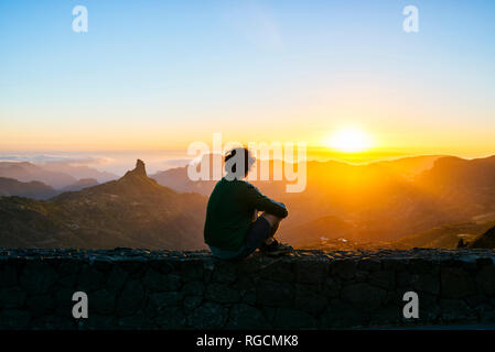 Espagne, Canaries, Gran Canaria, vue de dos de l'homme assis sur un mur en regardant le coucher de soleil sur mountainscape Banque D'Images