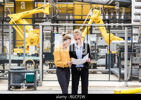 Deux femmes discuter plan en magasin d'usine étage avec robot industriel Banque D'Images