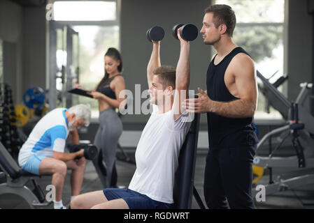 La formation de personnes dans une salle de sport moderne, à l'aide de l'équipement sportif professionnel. Formateur musculaire à la recherche au client. T shirt Homme en blanc assis avec les mains en l'air, holding dumbbells, l'élaboration. Banque D'Images