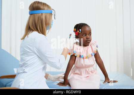 Vue rapprochée des cultures de petite fille et femme. Petite fille assise sur le lit pendant la consultation médicale. Jeune médecin en uniforme assis à la tenue à l'écart de l'équipement médical et aider les fille. Banque D'Images