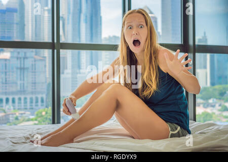 Jeune femme nitae sur le lit chez lui et faire l'épilation avec l'épilateur sur les jambes et est dans la douleur. Sur l'arrière-plan d'une fenêtre donnant sur la grande ville Banque D'Images