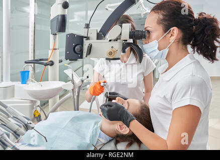 Vue de côté femme dentiste dans processus de travail en cabinet dentaire. Femme en uniforme blanc et masque à la recherche au microscope et de mettre l'implant. Patient couché sur le fauteuil dentaire et les soins sur les dents. Banque D'Images