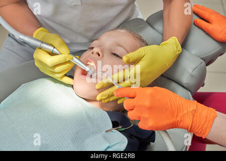 Petite fille couchée sur dentiste président et avoir fait les dents. Les dentistes de traiter et de guérir les dents avec des instruments spéciaux et de l'équipement. Les médecins professionnels portant des gants jaunes et orange. Banque D'Images