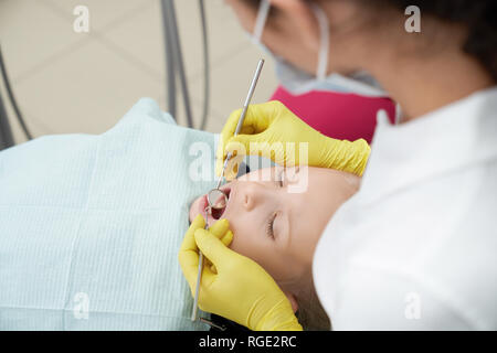 Portrait of cute girl lying on fauteuil dentaire avec la bouche ouverte tandis que les femmes médecin examinant les dents. Petit enfant médecin de passage en clinique. Concept de médecine dentaire et de stomatologie. Banque D'Images