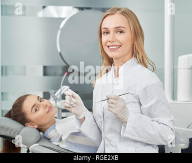 Belle femme, médecin de médecine dentaire portant en sarrau blanc, des gants qui posent avec patient. Charmante blonde smiling, looking at camera, tenant les instruments. Couché sur Client dentiste président. Banque D'Images