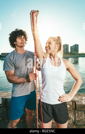 Jeune couple de remise en forme est en train de faire l'entraînement avec bande en caoutchouc par la rivière dans un coucher du soleil. La femme s'étire les bras et l'homme l'appuie. Banque D'Images
