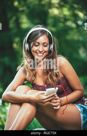 Femme se détend avec un casque d'écouter de la musique assis dans un parc. Elle les sms sont des messages sur son smartphone Banque D'Images