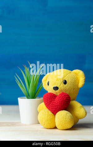 Faire part coeur rouge fil mis sur l'ours en peluche jaune en face de pot blanc et vert plantes ornementales sur table en bois et fond bleu. Concept de va Banque D'Images