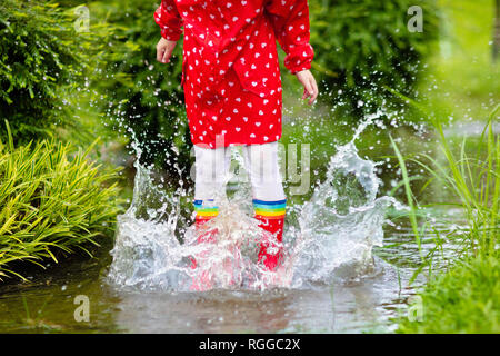 Kid jouer sous la pluie en automne parc. Le saut de l'enfant dans la boue flaque sur l'automne des pluies 24. Petite fille dans des bottes de pluie et veste rouge à l'extérieur par temps sh Banque D'Images