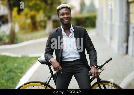 Young African man smiling avec earbeans lister voix de son portable tout en se tenant sur sa bicyclette sur une rue de la ville Banque D'Images