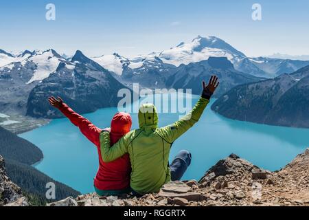 Vue depuis le sentier de randonnée Panorama Ridge, deux randonneurs assis sur un rocher qui s'étend les bras en l'air, Garibaldi Lake, turquoise Banque D'Images