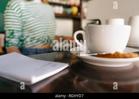 Tasse de café et un bloc-notes sur la table dans un café avec woman in background Banque D'Images