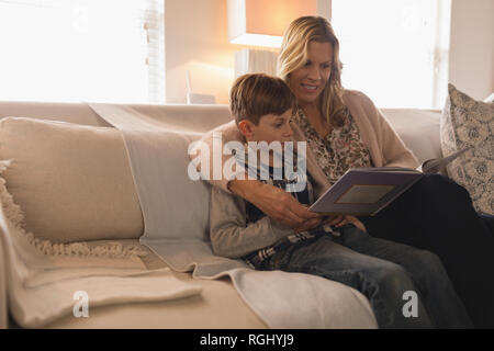 Vue latérale de la mère avec son fils de passer du temps ensemble tout en lisant un livre dans la salle de séjour à la maison Banque D'Images