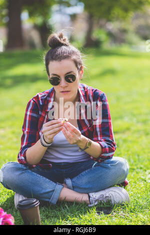 Vue de face d'une jeune femme de taille basse assise sur l'herbe dans un parc tout en tenant une fleur par une journée ensoleillée Banque D'Images