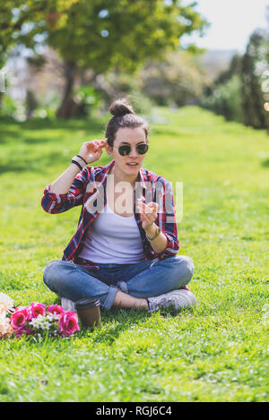 Vue de face d'une jeune femme de taille basse assise sur l'herbe dans un parc tout en tenant une fleur par une journée ensoleillée Banque D'Images