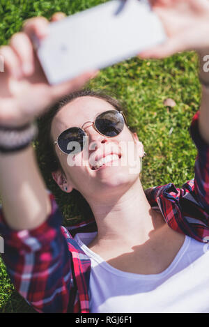 Au-dessus de la vue d'une jeune fille hipster souriante couché sur l'herbe dans une journée ensoleillée dans un parc tout en prenant un selfie avec un téléphone mobile Banque D'Images